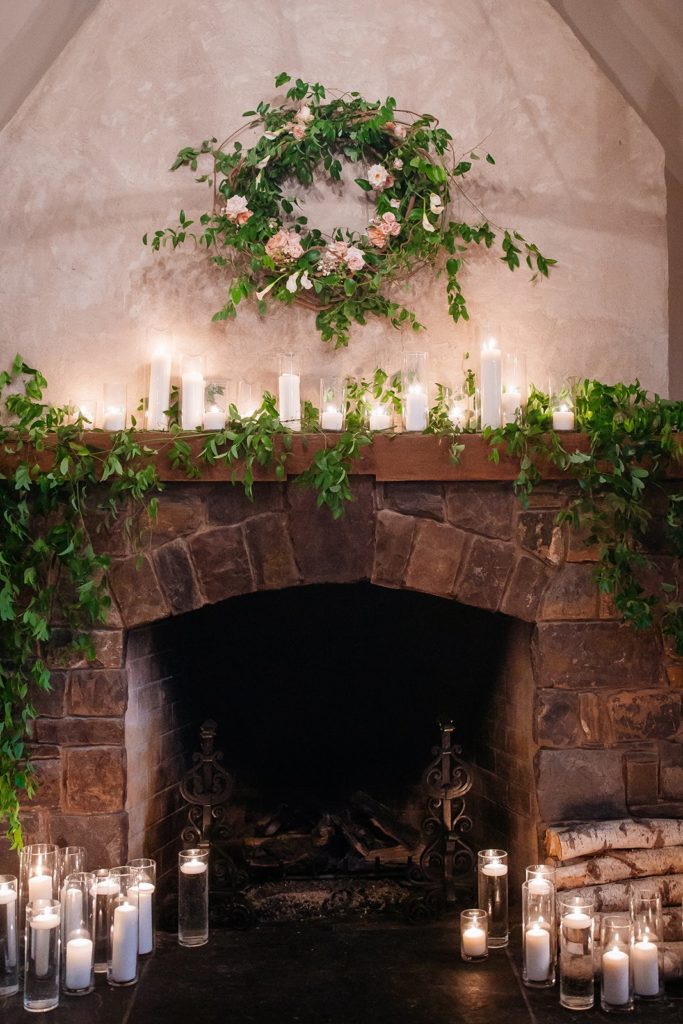 Elegant Secret Garden Wedding Reception at Barley Sheaf Farm, Fireplace decorated with multiple pillar candles enclosed in glass in varying heights as well as water-filled vases topped with floating candles. Finished with a beautiful garden-inspired wreath on the wall above featuring lush greenery and accents of flowers in ivory and peach. Designed by Sebesta Design. Photography by Juliana Laury Photography