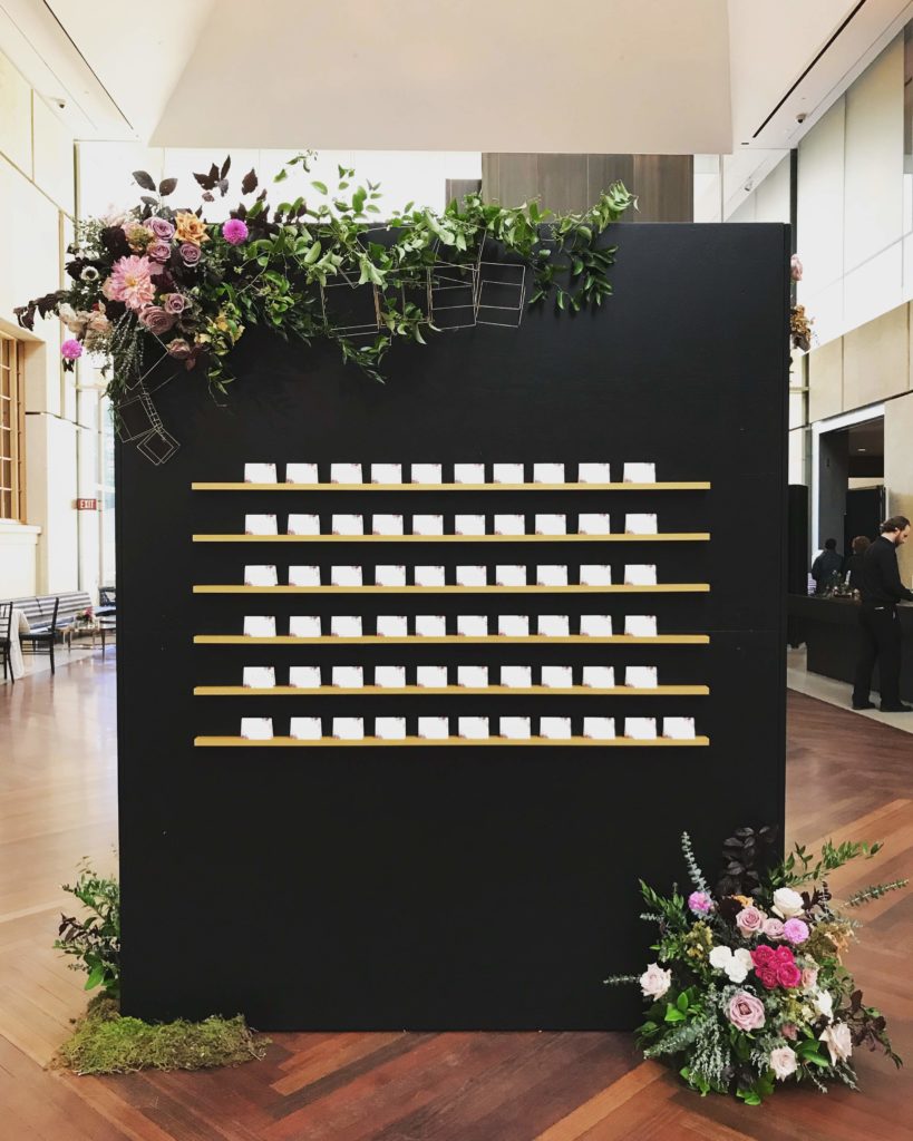 Custom Museum Wedding Escort Card Wall Display, striking black background with asymmetrical floral details including dahlias, garden roses, wild smilax and gold geometric wire forms at the Barnes Foundation by Sebesta Design. Photography by Max Grudzinski Photography 