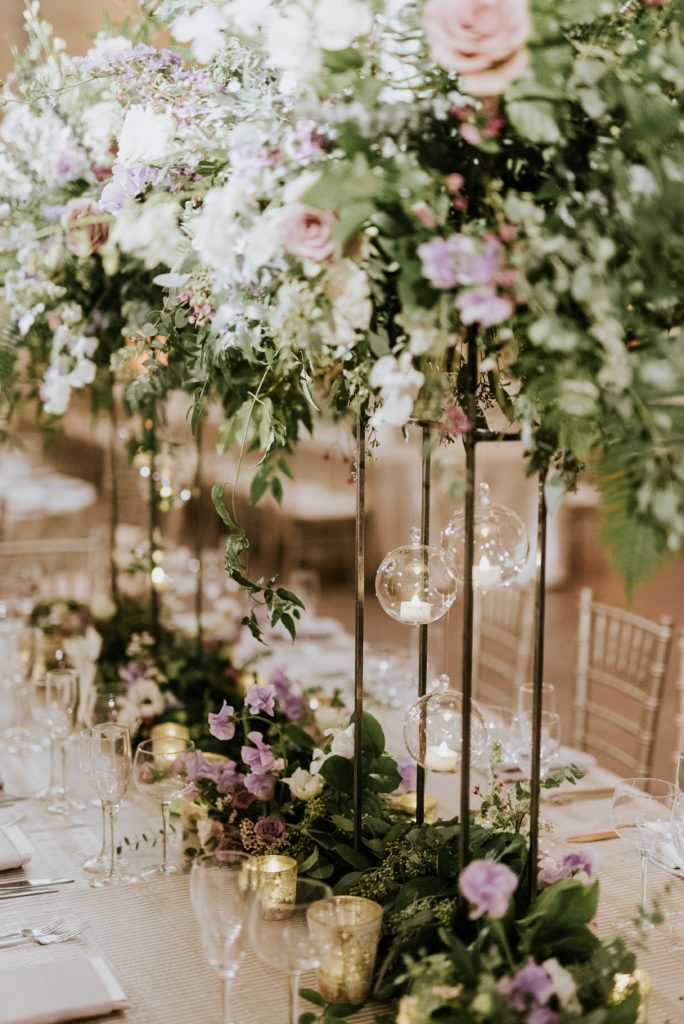 Winter wedding centerpiece featuring roses, larkspur, delphinium, fern, eucalyptus, fern, jasmine vine and ruscus on tall metal stands with hanging candles at the Barnes Foundation. Photo by M2 Photography.