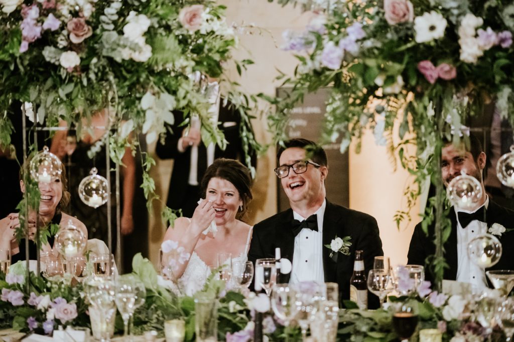 Bride & groom framed by tall centerpieces, hanging candles, vine and flowers on their King's table at their New Year's Eve wedding at the Barnes Foundation.