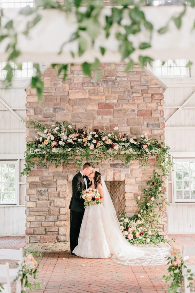 Bride & Groom standing in front of the fireplace embellished with flowers and foliage at the Ashford Estate by Sebesta Design. Photo by Emily Wren Photography.