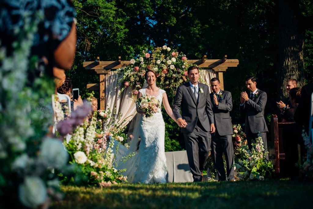 Newly Married Couple strolling back up their beautiful garden-inspired aisle following their outdoor wedding ceremonyat The Inn at Barley Sheaf Farm. Decorated floral and draped fabric arbor in the background. Designed by Sebesta Design. Photography by Juliana Laury Photography