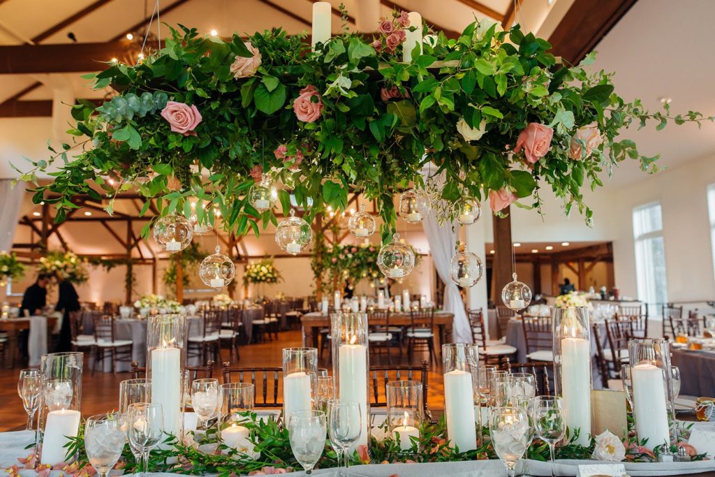 Elegant Secret Garden Wedding Reception, Hanging Floral Centerpiece with draping greenery, accents of peach, ivory and coral flowers and hanging candles above and multiple types of candles in varying sizes and shapes below with accents of greenery and blooms to create a runner effect down a long table at The Inn at Barley Sheaf Farm. Designed by Sebesta Design. Photography by Juliana Laury Photography