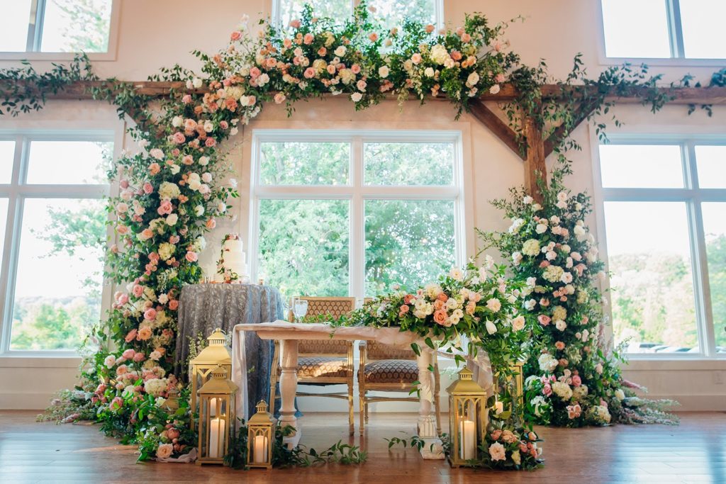 Dramatic sweetheart table backdrop filled with flowers and foliage that wraps from the floor to wall behind it completely encompassing the sweetheart table at The Barley Sheaf Farm by Sebesta Design. Photo by Juliana Laury Photography.