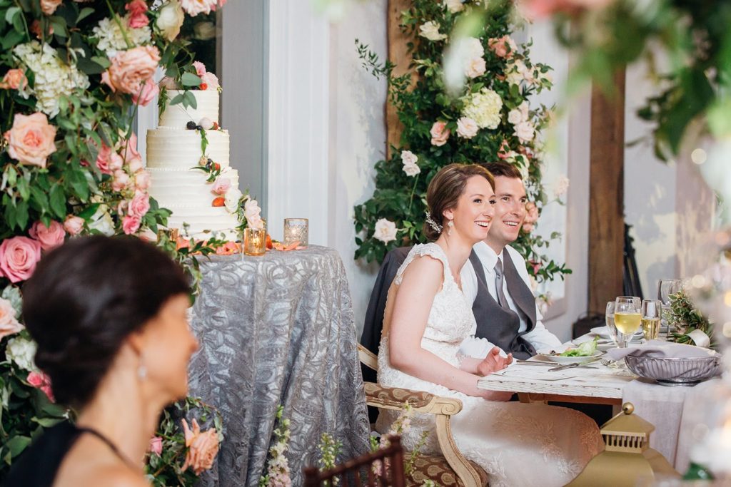 Bride and Groom at their sweetheart table, enclosed in a secret garden-inspired immersive floral installation with abundant greenery and flowers in shades of ivory, peach and coral. The Inn at Barley Sheaf Farm wedding reception designed by Sebesta Design. Photography by Juliana Laury Photography