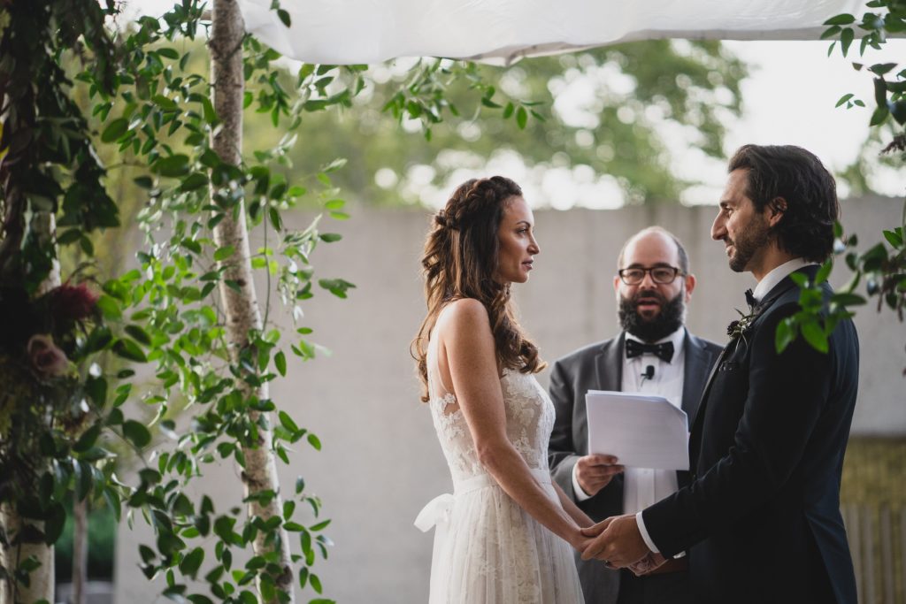 Bride and Groom reciting vows during Modern Organic Museum Wedding outdoor Ceremony under organic smilax covered chuppah at the Barnes Foundation by Sebesta Design. Photography by Max Grudzinski Photography 