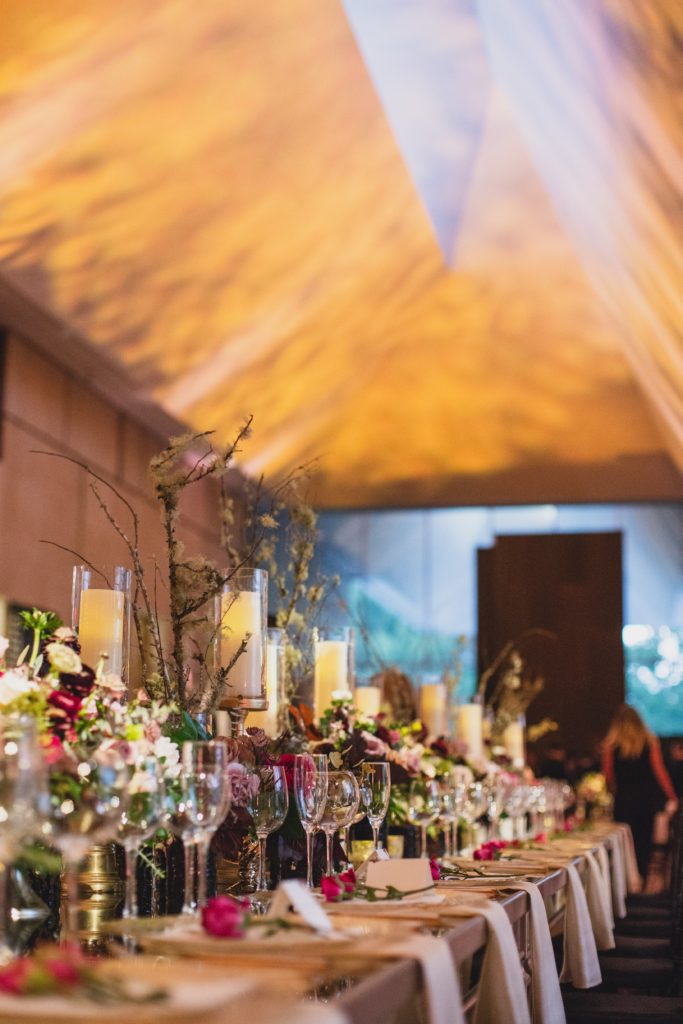 Detail of Striking Museum Wedding Reception long head table centerpiece design using multiple types of candles in varying heights, organic textural arrangements of flowers and foliage, including natural elements such as branches all down a mirrored table at the Barnes Foundation by Sebesta Design. Photography by Max Grudzinski Photography 