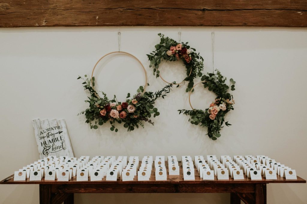 Romantic Unique Escort Card Display at The Inn at Barley Sheaf Farm featuring three lush floral hoops suspended over the long wood farm table lined with custom escort cards featuring wax seals and calligraphy for an old world feel. Designed by Sebesta Design. Photography by M2 Photography.