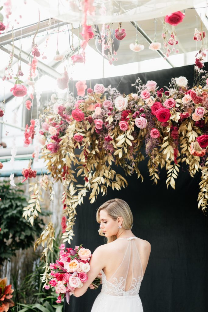 Love is in the Air styled shoot and bespoke bridal event at the Horticulture Center, custom photo backdrop with lush hanging floral piece in shades of pink, accented with gold painted foliage against a dramatic black backdrop. Model wearing Limor Rosen and carrying a beautiful pink bridal bouquet with sweet peas, roses and garden roses . Event design and florals by Sebesta Design. Photography by Emily Wren Photography. 