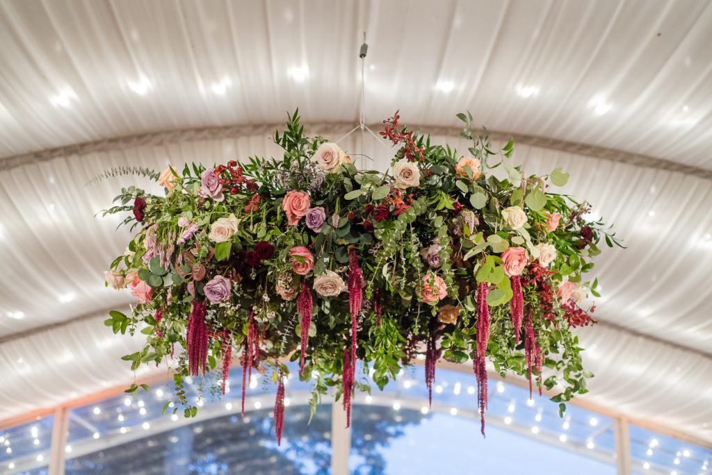 Bold Boho Organic Wedding Reception custom floral hanging chandelier featuring wild smilax vine, quicksand roses, spray roses, peach shimmer roses, mauve roses, blush stock, antique coral roses and hanging amaranthus and garden greens designed by Sebesta Design at Glen Foerd on the Delaware. Photographed by The Caffeinated Photographer.